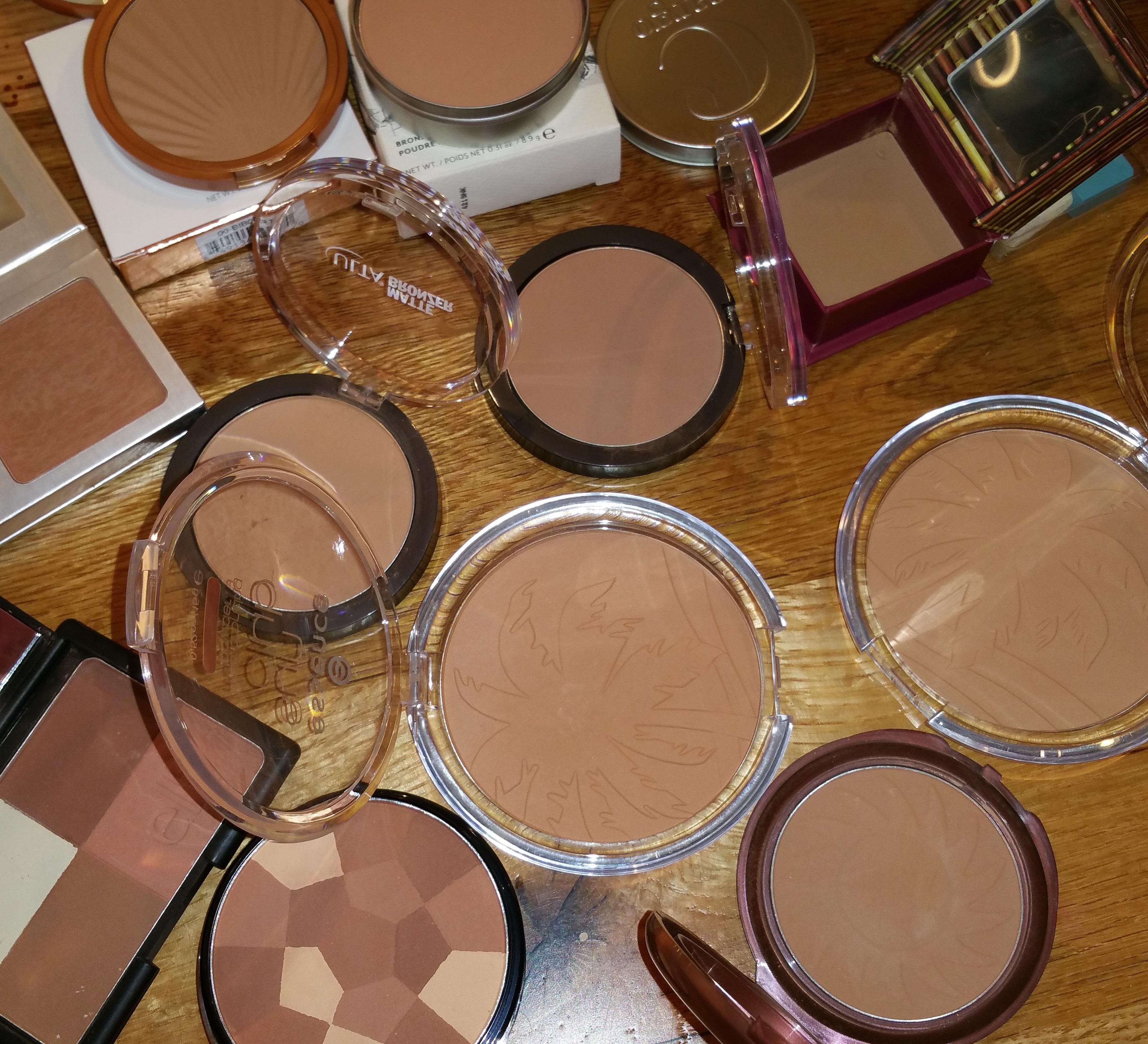 Starting from bottom left: e.l.f. Cool Bronzer, NYC Color Wheel - All Over Bronze, and Smooth Skin Bronzing Face Powder; Second row right to left: Essence Sun Club - 01 Natural and 02 Sunny; Third row: left to right: Ulta Matte Bronzer Warm and Cool; Top row right to left: Benefit Hoola, Cargo Matte Bronzer - Medium, Laura Geller Matte - Medium, and It Cosmetics CC + Bronzer.